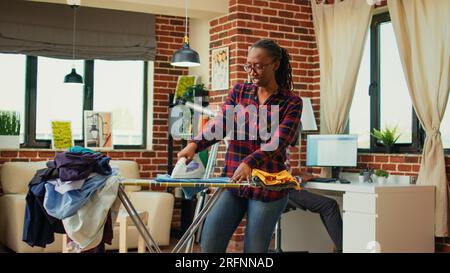 Female adult enjoying doing laundry work at home, having fun listening to music and ironing washed clothes. Happy housewife dancing and singing while she does chores, iron with steam. Stock Photo