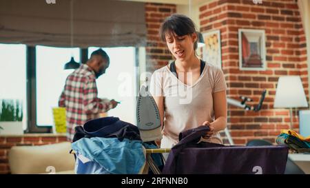 Happy adult singing and dancing while she irons clothes, housewife having fun spring cleaning in living room. Cheerful woman enjoying ironing garments and listening to music. Handheld shot. Stock Photo