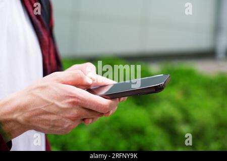 Unrecognizable man in red shirt using a modern mobile smartphone in the hands on the outdoors, 5g internet, text messages, video calls Stock Photo