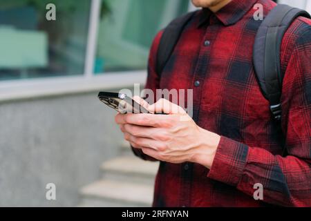 Unrecognizable young modern urban man in red shirt using modern mobile smartphone in hands-on outdoors, 5g internet, using text messages, video calls Stock Photo