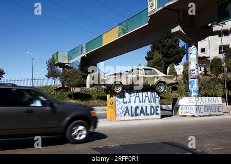 Car driving past crashed car on pedestal in middle of motorway warning drivers not to drink drive and drive carefully, La Ceja, El Alto, Bolivia Stock Photo