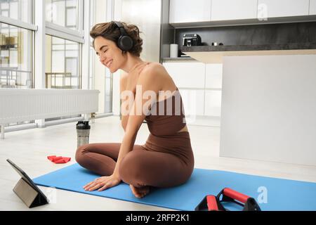 Smiling brunette fitness girl, listens music in headphones, watches online video from tablet, learns yoga poses, workout tips, does pilates exercises Stock Photo