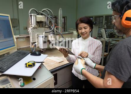 Asian female company manager showing work on digital tablet to CNC machine operator in factory. Stock Photo