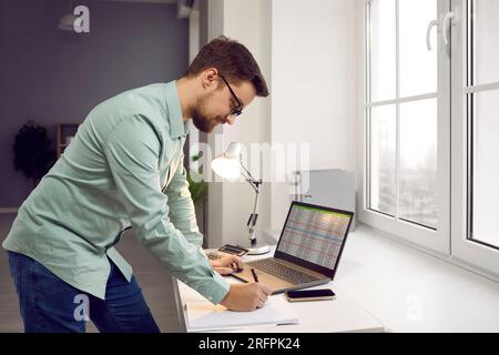 Focused man standing at desk working on laptop and writing in notebook Stock Photo