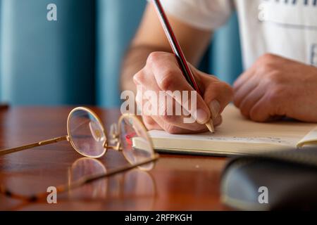 A detail of a writer with his glasses and pencil writing in a paper. Taking notes with his pencil on a notebook with his glasses laying down. Stock Photo