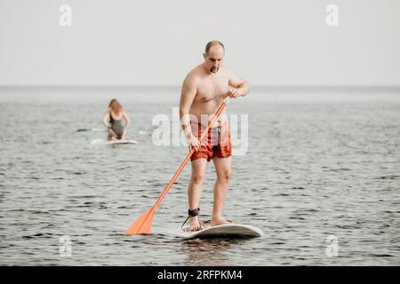 Active mature male paddler with his paddleboard and paddle on a sea at summer. Happy senior man stands with a SUP board. Stand up paddle boarding - Stock Photo