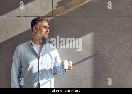 Gray haired bearded middle aged man standing at gray wall with coffee mug Stock Photo