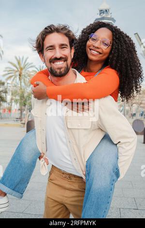 Verical portrait of couple embracing and having fun. Man giving his african american girlfriend a romantic piggyback ride enjoying a nice day together. Smiling guy in love carrying on back his woman. High quality photo Stock Photo