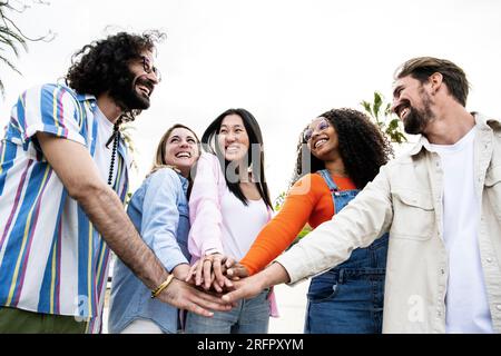 Diverse group of cheerful millennial friends standing together stacking hands in the street. Cheerful multiethnic young carefree friends laughing and Stock Photo