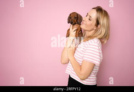 Young happy woman hugging and kissing her dachshund puppy dog isolated over pink background. Stock Photo