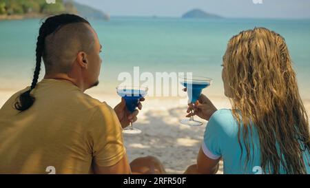 The love couple is holding a glass of blue curacao cocktail, on sea. Man with plait and blonde woman drink alcohol on sand beach in shadow by azure ocean of a tropical island. Summer holiday concept. Stock Photo