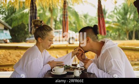 Attractive young man and woman eat and drink tasty beverages spending time in traditional local floating cafe on water. Loving happy couple having breakfast outdoors, feed each other. Tropical view Stock Photo