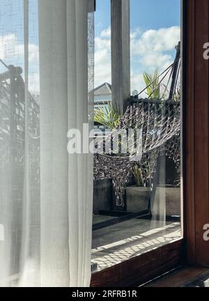 Comfortable boho style grey macrame hammock and palm houseplants on balcony. View from room. Stock Photo