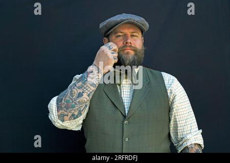 Bearded man is twirling his mustache with his fingers. Portrait of a hipster on black background closeup male with tattoos on his arm. Stock Photo