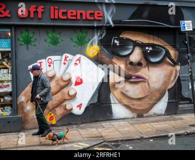 Brighton UK - Man with small dog walks past a Glimmer Twins mural of cigar smoking card player in Brighton Stock Photo