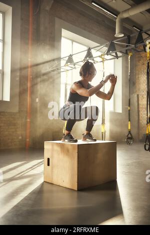 Challenging myself. Side view of athletic woman in sportswear doing squat while standing on wooden box at gym Stock Photo