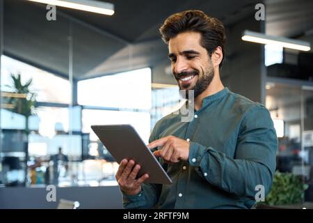 Happy young Latin business man using digital tablet standing in office. Stock Photo