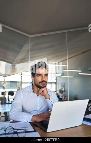 Thoughtful young Latin business man looking away thinking using laptop at work. Stock Photo