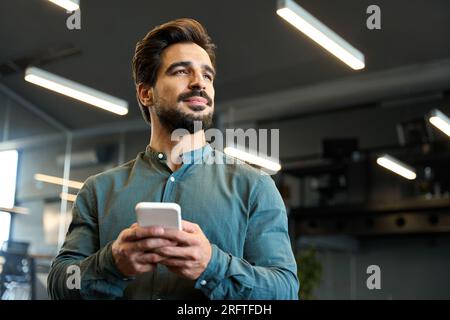Young Latin business man entrepreneur holding mobile phone standing in office. Stock Photo