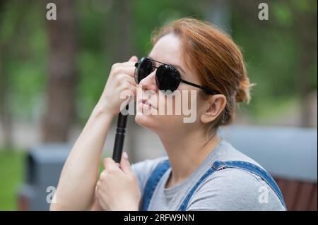 Blind red-haired woman sitting outdoors leaning on a cane.  Stock Photo
