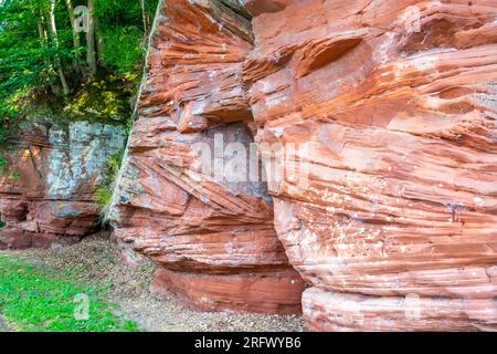 Red sandstone rocky slope, irregular texture, mold, white spots and linear erosions caused by passage of time, trees with green foliage in background, Stock Photo