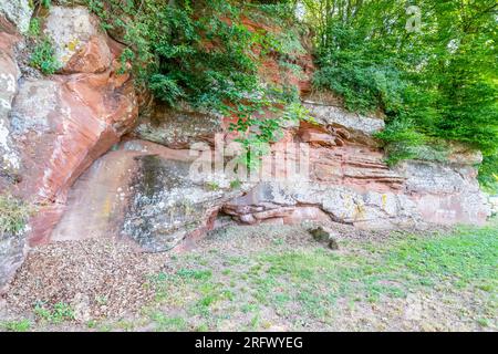 Rocky slope of red sandstone between sparse grass and green foliage in background, unevenly textured wall, mold, white spots and linear erosions, sunn Stock Photo