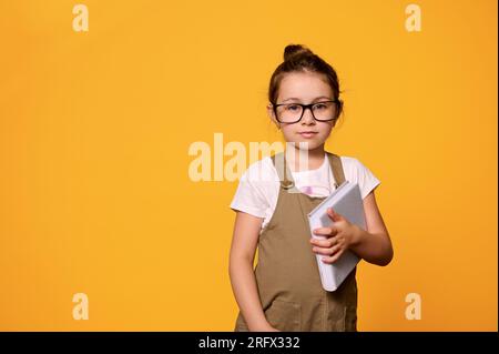 Adevrtising studio portrait of Caucasian confident serious cute little girl 6 years old, holding textbook, dressed in casual clothes and eyeglasses Stock Photo
