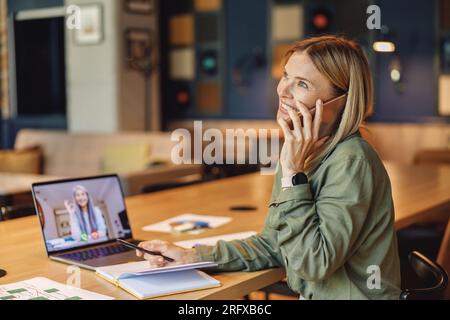 Woman entrepreneur talking phone during video conference with colleague while sitting in cafe Stock Photo