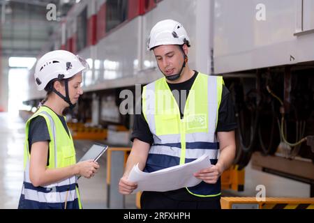 Young caucasian engineer man and woman or worker look blueprint and checking electric train for planning maintenance in station, transport and infrast Stock Photo