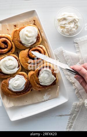 A hand spreading frosting on freshly baked cinnamon buns. Stock Photo