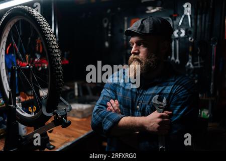 Portrait of thoughtful bearded repairman in cap holding wrench standing with crossed arms by bicycle in repair workshop with dark interior, looking Stock Photo