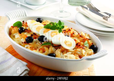 Baked cod with onion, black olives, boiled eggs and oregano. In an oval iron baking dish. perpendicular view. Stock Photo