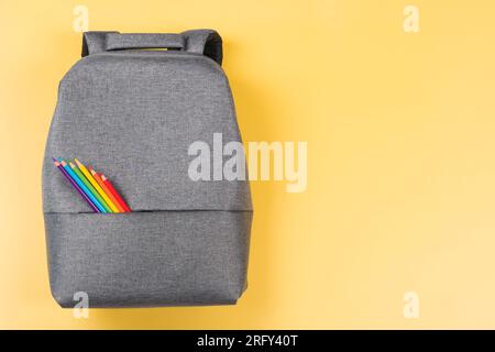 Gray backpack with colored pencils in rainbow colors on yellow background. Top view, copy space. Back to school, education, learning concept Stock Photo