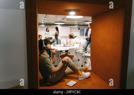 Male employee listening to music on his headphones and over-thinking while his diverse colleagues are working in the back Stock Photo
