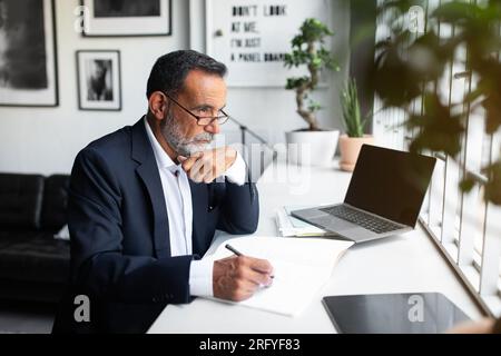 Pensive serious caucasian old businessman in suit with contract make notes, look at computer with empty screen Stock Photo