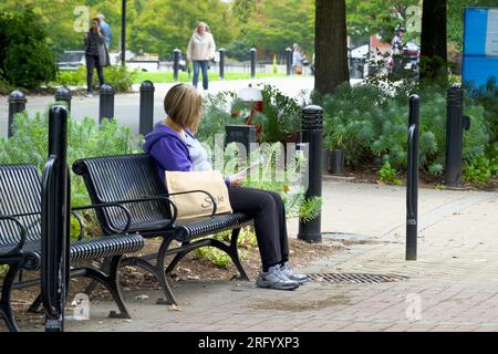 Young blonde woman sitting on a black, slatted park bench with a brown shopping bag reading a magazine - Lower Mainland, B. C., Canada.  - Sept. 2019. Stock Photo
