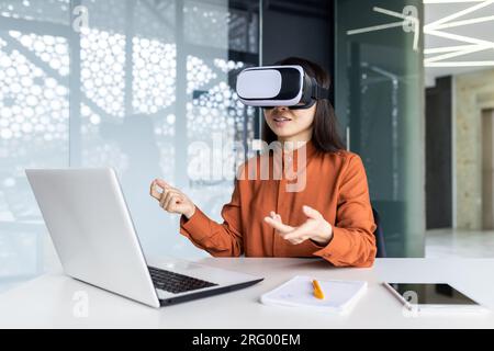 Asian woman wearing virtual reality glasses sitting at a desk in a workspace inside the office, businesswoman in online meeting talking remotely with colleagues, virtual meeting simulation. Stock Photo