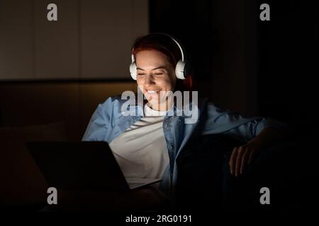 Relaxed carefree female sits comfortably with computer wear headphones watching funny movie at night Stock Photo