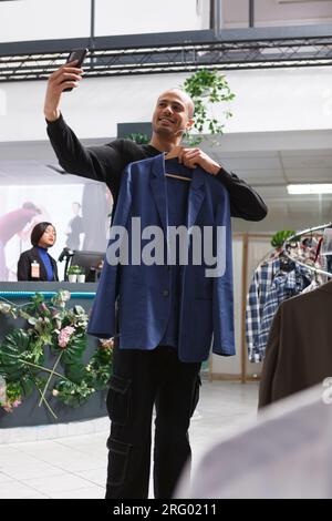 Clothing store arab man client holding jacket on hanger and taking photo on smartphone while browsing apparel in shopping mall. Boutique smiling buyer chatting in videocall and showing outfit Stock Photo