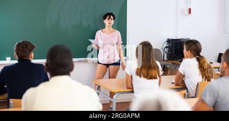 Young teacher explaining new topic while standing at blackboard Stock Photo