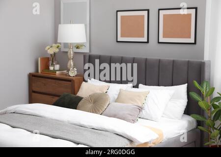 Comfortable bed with cushions, lamp and different decor on wooden chest of drawers in room. Stylish interior Stock Photo