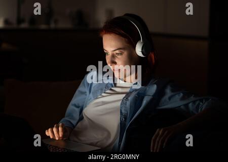 Concentrated middle aged programmer female sits on chair work on laptop at night. Freelancr concept Stock Photo