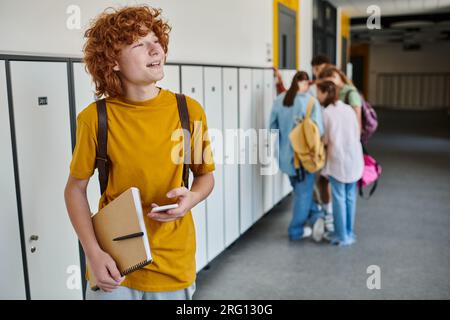cheerful redhead schoolboy using smartphone and holding notebook, blur, students in school hallway Stock Photo