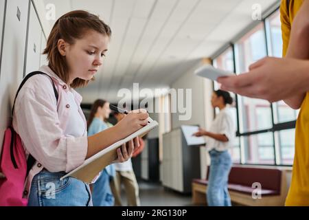 focused schoolgirl taking notes, holding notebook near boy using smartphone in school hallway Stock Photo