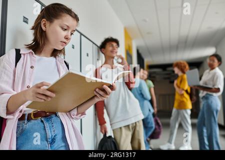 schoolgirl holding notebook and standing near blurred and happy teen classmate, students, hallway Stock Photo