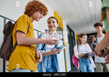 back to school, classmates using gadgets in school hallway, redhead boy using smartphone near girl Stock Photo