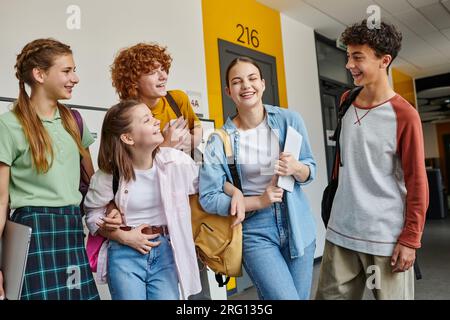 teenage schoolkids laughing and standing in school hallway together, teen classmates with devices Stock Photo