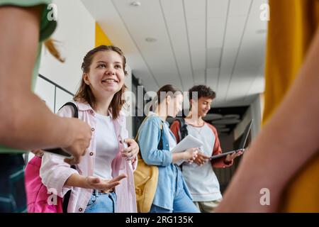 back to school, happy teen girl talking to classmates in hallway, blurred students using devices Stock Photo