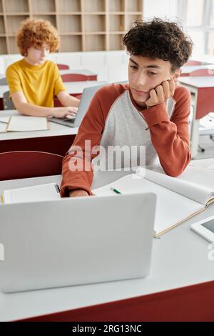 bored curly boy using laptop in classroom, back to school concept, e-learn, assignment, classmates Stock Photo