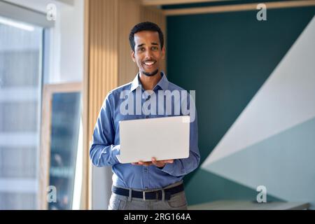 Smiling young business man standing in office with laptop, portrait. Stock Photo
